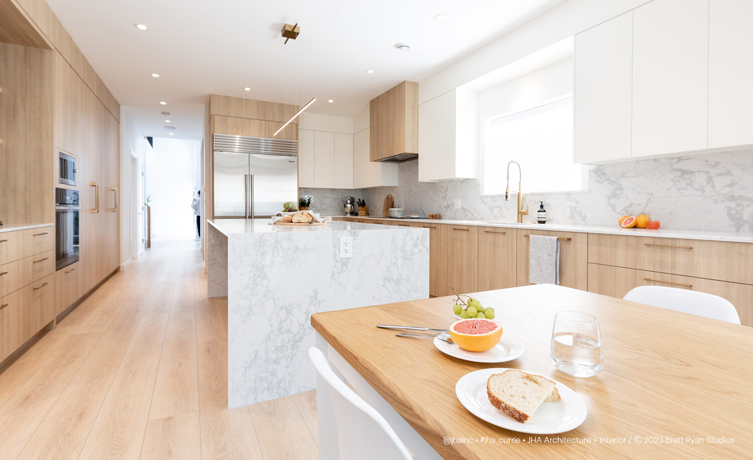 Photograph of bright and open kitchen, with wood flooring and cabinetry, and white marble counter accents